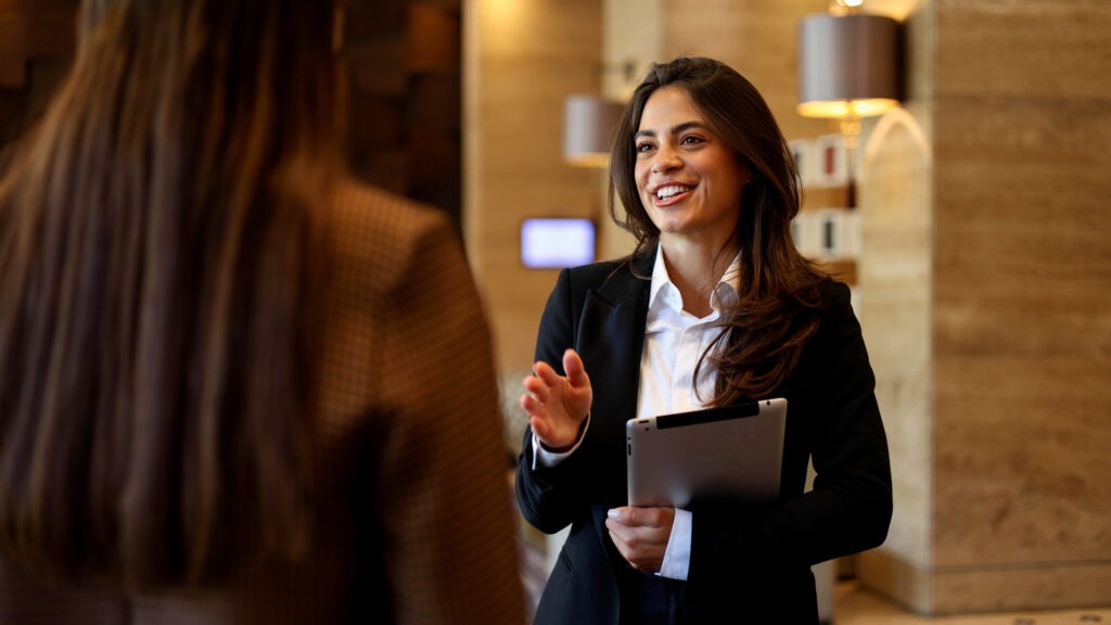 Businesswoman talking with colleague, holding digital tablet in modern office or hotel lobby, smiling during discussion